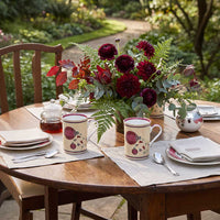 A styled outdoor dining table set with cream-colored ceramic mugs decorated with red berry and floral motifs, alongside a matching floral centerpiece, teapot, and plates, set against a lush garden background.