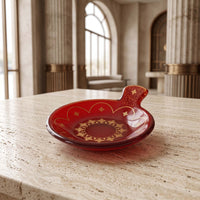 A red glass dish with a small handle and gold decorative patterns, set on a light stone table in a grand, softly blurred interior with columns and arched windows.