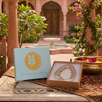 Decorative plate in a box with a decorative box featuring a logo and website text, set against an ornate garden background.