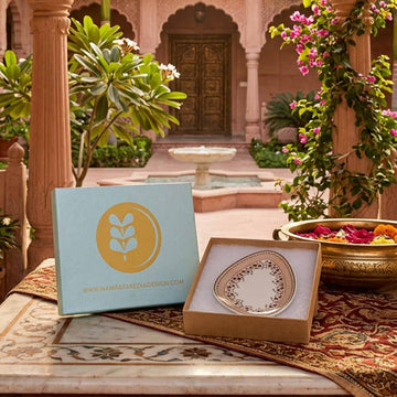 Decorative plate in a box with a decorative box featuring a logo and website text, set against an ornate garden background.