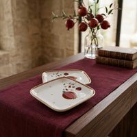 Two square ceramic plates with rounded corners, decorated with a red strawberry motif and gold trim, arranged on a dark burgundy table runner over a wooden table in a warm, rustic interior setting.
