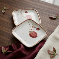 Two square white ceramic plates with rounded corners, decorated with red pomegranate motifs, gold dots, and gold-rimmed edges, arranged on a dark wooden table with draped burgundy fabric and scattered leaves.