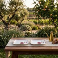 Two square white ceramic plates with red and gold floral details are set on a wooden table outdoors in a sunny garden, alongside a gold teapot and grapes.