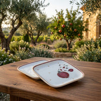 Two square white ceramic serving plates with red fruit illustrations and gold trim, stacked on a wooden table in an outdoor garden setting.