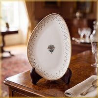 An oval white porcelain serving platter with a gold rim, delicate black dotted border pattern, and a central floral motif, displayed on a dark wooden stand on a polished wooden table.