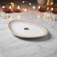 An oval white serving platter with a gold rim and a small central floral motif, styled on a marble surface with lit candles and white flowers in the background.