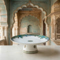 A white ceramic pedestal plate with intricate blue and green floral patterns, displayed on a light stone surface with a blurred architectural background.