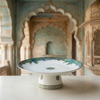 A white ceramic pedestal plate with intricate blue and green floral patterns, displayed on a light stone surface with a blurred architectural background.