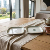 Two square white ceramic plates with a green and blue patterned border and a small central medallion, displayed on a wooden table runner in a bright dining room.