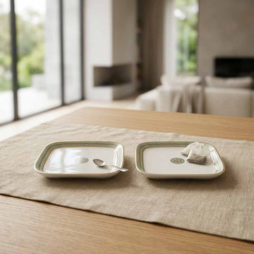 Two square white ceramic plates with green patterned borders are arranged on a beige table runner on a wooden table, with a teacup and a small teabag packet on top. The background shows a softly blurred modern living room interior.