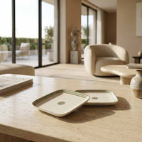 Two square ceramic coasters with rounded corners, featuring a green and gold patterned border and a circular green medallion in the center, displayed on a beige stone table in a modern, sunlit living room.