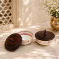 Two ceramic bowls with wooden lids on a textured surface with a decorative vase in the background.
