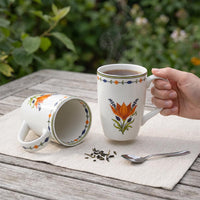 A white ceramic mug with a colorful orange and green floral design is shown on a wooden table outdoors, with one mug lying on its side and a hand holding the other mug. Steam rises from the mug, and dried tea leaves and a spoon are placed nearby on a beige cloth with a blurred garden background.