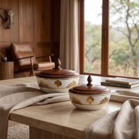 Two ceramic lidded bowls with dark wood lids and gold tree motifs, displayed on a stone table in a rustic interior setting.