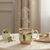 Steaming mug with a nature-themed design on a table with a plant and fabric in the background.