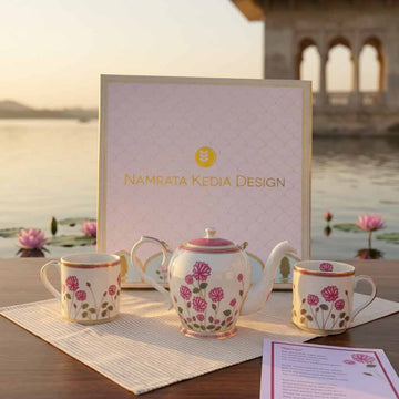Tea set with floral design on a table with a scenic background, featuring 'Namrata Kedia Design' branding.