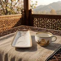 A white ceramic rectangular plate with gold decorative borders and a matching white ceramic cup with gold motifs, set on a beige linen cloth on a carved wooden table with a scenic outdoor background.