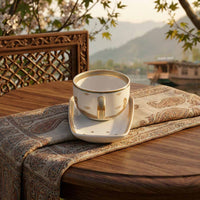 A white ceramic teacup and cookie plate set with gold detailing, placed on a patterned runner on a wooden table with a scenic outdoor background.