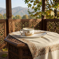 A white ceramic teacup and saucer with gold detailing sits on a patterned cream tablecloth draped over a carved wooden table, with a scenic mountain background visible through a wooden gazebo.
