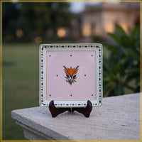 A square decorative ceramic tile with a light pink center, featuring an orange tulip motif and a patterned border, displayed on a small dark wooden stand outdoors on a stone ledge with a blurred garden background.