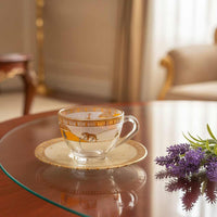 Decorative glass teacup with gold accents on a saucer, placed on a glass table with a blurred indoor background.