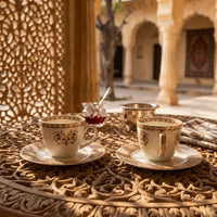Two white ceramic teacups with gold and brown floral patterns on saucers, set on an ornate carved wooden table in a sunlit courtyard with arches and columns in the background.