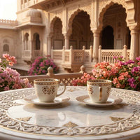 Two ornate white teacups with gold detailing on saucers, set on a marble table in front of a traditional Indian courtyard with pink flowers.