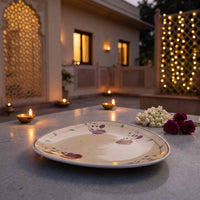 A round ceramic plate with a pomegranate pattern, displayed on a stone table in an outdoor evening setting with lit candles, flowers, and warm background lights.