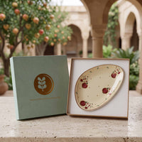 An oval ceramic plate with a cream background and red pomegranate motifs, displayed in an open presentation box next to its light green branded outer box on a stone surface, with a blurred courtyard background.