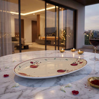 An oval ceramic serving platter with a pomegranate pattern, set on a white marble table in a modern, elegantly lit dining room with a glass of red wine and candles nearby.