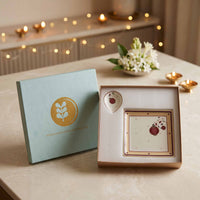 A boxed tea or matcha gift set displayed on a tabletop, with a light blue outer box and an open inner box showing a square cream-colored tea packet or card with red floral motifs. The scene is softly lit with candles and warm string lights in the background.