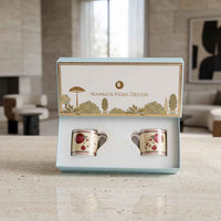 A boxed set of two small ceramic cups with a floral print, displayed open in a light blue presentation box on a countertop. The box lid is printed with the brand name 'Namrata Kedia Design' and decorative landscape artwork.