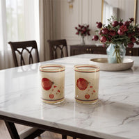 Two decorative glasses with gold trim and red strawberry illustrations sit on a white marble dining table in a beautifully lit room with dark wood chairs and floral arrangements in the background.
