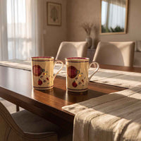 Two ceramic mugs with a cream base and red floral patterns sit on a wooden dining table in a softly lit home interior.