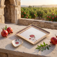 A square white ceramic serving platter and a small matching bowl with gold trim and a pomegranate illustration, styled on a stone ledge outdoors with pomegranates and a garden in the background.