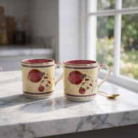 Two cream-colored ceramic mugs with red pomegranate and floral designs, sitting on a white marble countertop in a kitchen setting with a window in the background.