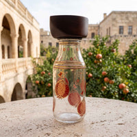 A clear glass bottle with a dark brown wooden lid, decorated with red and gold pomegranate motifs, set on a stone table with a blurred courtyard background.