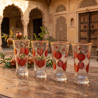 Four clear glass tumblers with red pomegranate motifs and gold rims, arranged on a wooden table in front of a traditional courtyard with arches and pomegranate branches.