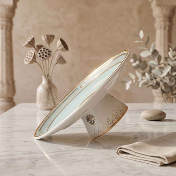 A ceramic pedestal plate with a light blue-green glazed interior and gold trim, displayed on a marble table in a styled interior setting with neutral decor.