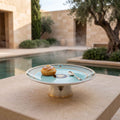 A round ceramic cake stand with a light aqua center and gold-trimmed rim sits on a stone terrace beside a pool, with a small pastry and spoon on top. The background shows a sunlit courtyard with stone walls, greenery, and a tree.