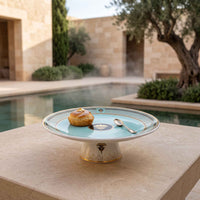 A round ceramic cake stand with a light aqua center and gold-trimmed rim sits on a stone terrace beside a pool, with a small pastry and spoon on top. The background shows a sunlit courtyard with stone walls, greenery, and a tree.