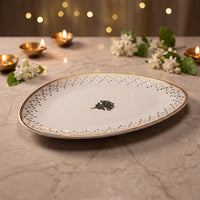 An oval white serving platter with a gold rim, delicate blue-green dotted border, and a central floral motif, displayed on a marble surface with warm bokeh lights and decorative flowers in the background.