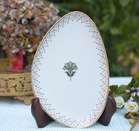 An oval white porcelain serving platter with a gold rim, delicate black dotted border pattern, and a central green lotus flower motif, displayed on a dark wooden stand.