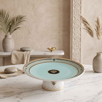A ceramic pedestal plate with a light aqua center, gold-trimmed rim, and white base, styled on a marble table in a warm neutral interior setting.