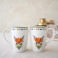 Two white ceramic mugs with orange floral designs and green leaves, set on a light textured surface with a soft, blurred background.