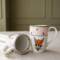 Two ceramic mugs with a hand-painted floral design, one standing upright and the other lying on its side to show the interior, set on a textured beige tablecloth with a soft-focus paneled wall background.