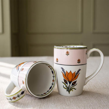 Two ceramic mugs with a hand-painted floral design, one standing upright and the other lying on its side to show the interior, set on a textured beige tablecloth with a soft-focus paneled wall background.
