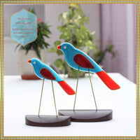 Two colorful bird figurines on stands with a blurred indoor background