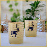 Two glasses  with a pale yellow tint and navy blue deer motifs, displayed on a white surface with a softly blurred background of green plants and decorative jars.