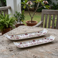 Two long rectangular ceramic serving trays with a white base, a dusty rose floral pattern, and gold trim, displayed on a stone table in an outdoor garden setting.